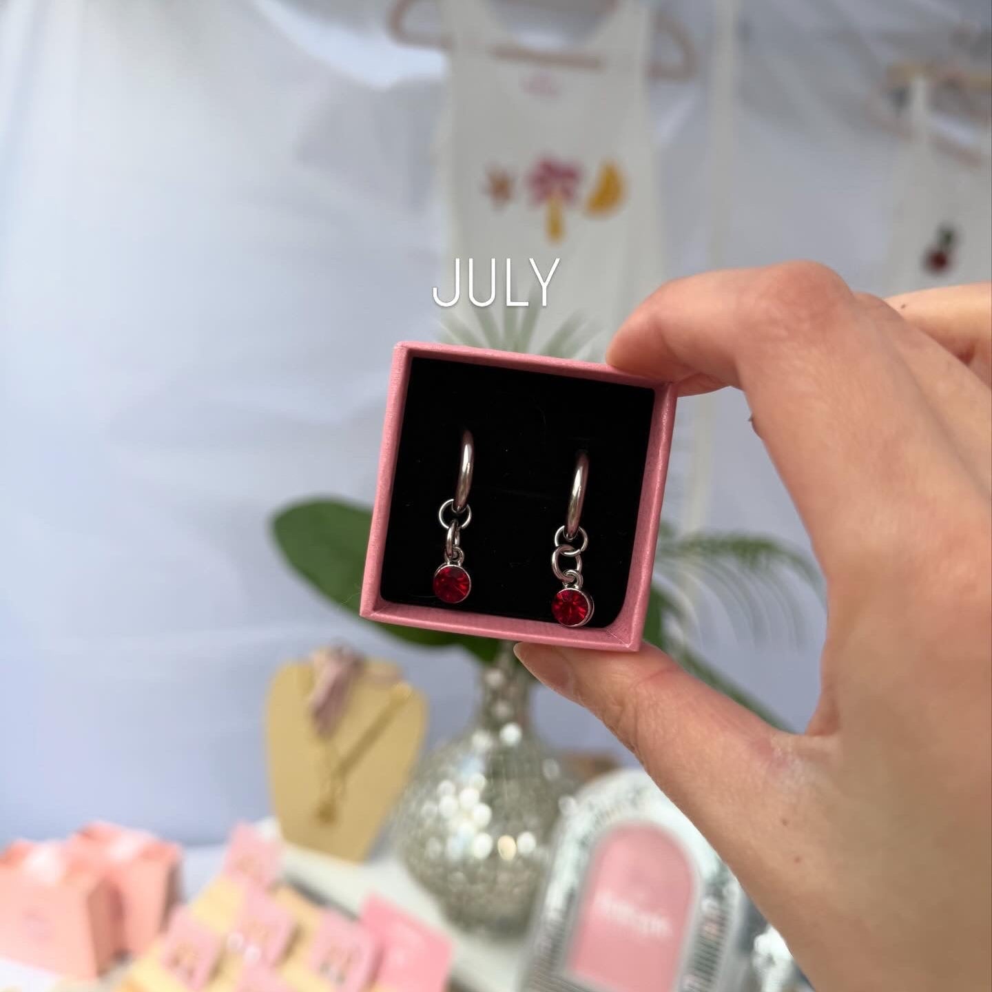 Pink box with earrings held by a hand, with 'JULY' text above.