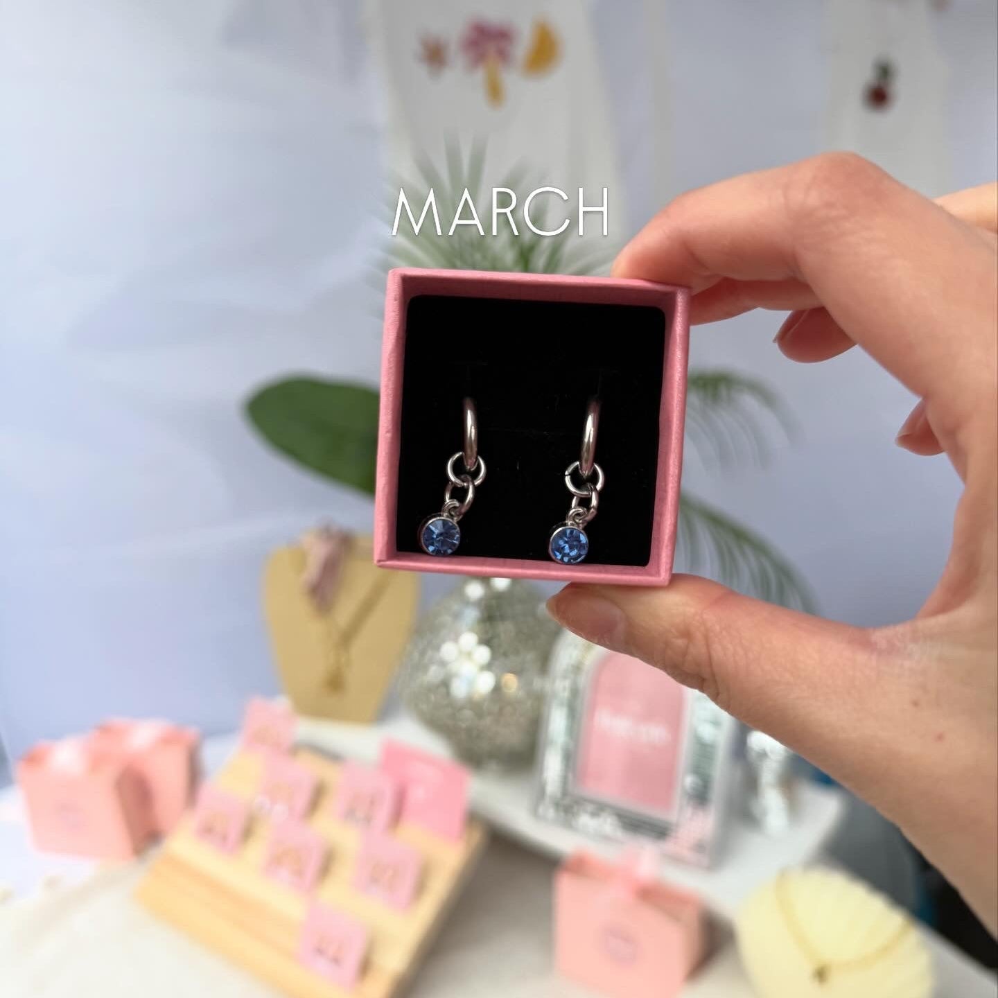Hand holding a box with earrings labeled 'MARCH' on a decorative background.