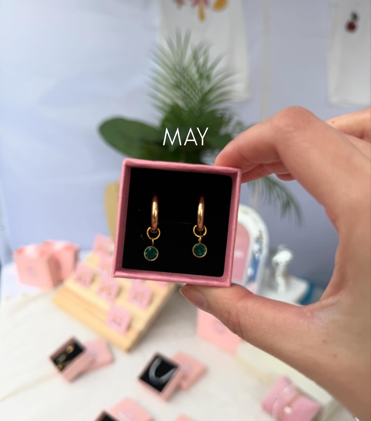 Gold earrings with green stones in a pink jewelry box held by a hand, with 'MAY' text above.
