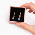 Gold earrings with blue stones in a pink box held by a hand on a white background