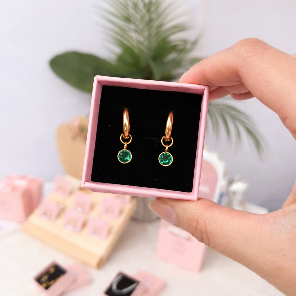 Gold earrings with green gemstones in a pink box held by a hand.