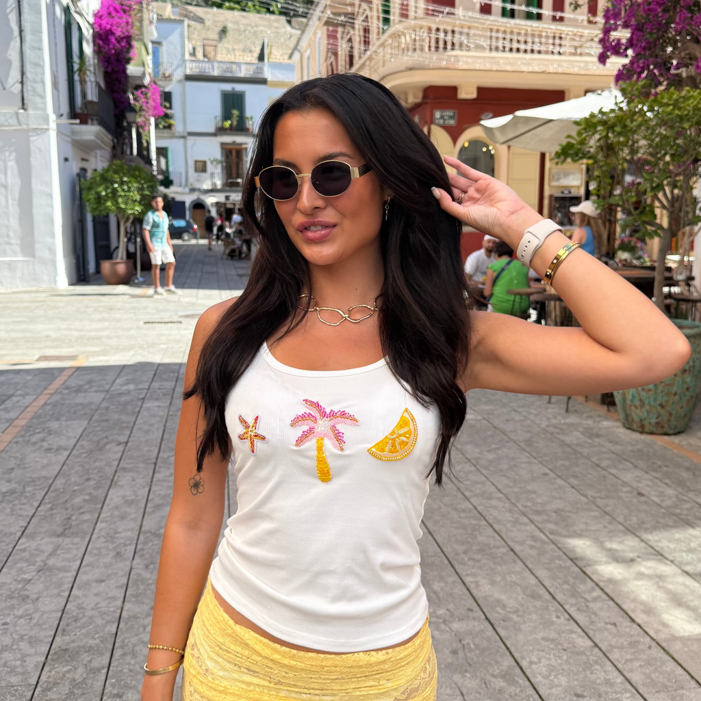 Woman in a white tank top with palm tree and sun design and yellow skirt standing on a street with buildings and flowers in the background.