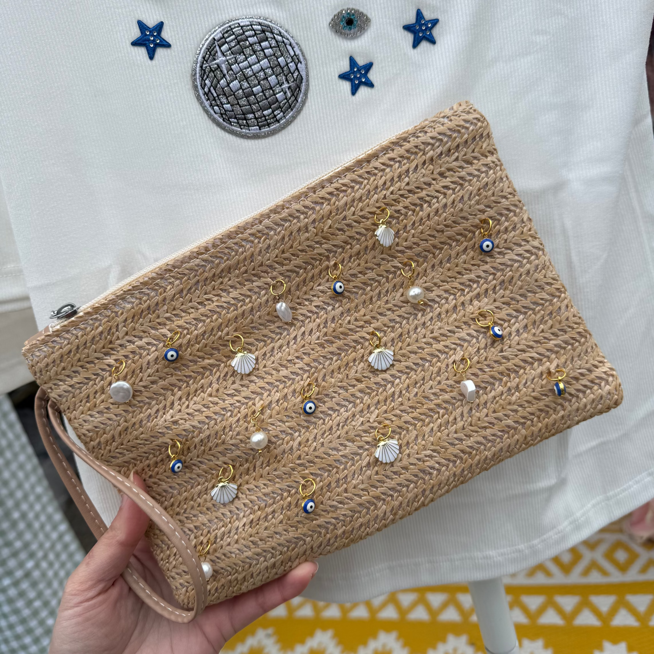 Beige clutch bag with decorative elements held by a hand against a white garment with colorful patches.